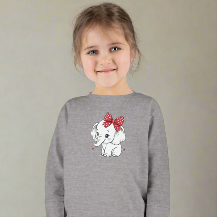 Toddler girl smiling in a gray long sleeve tee featuring a cute baby elephant with a red bow design.